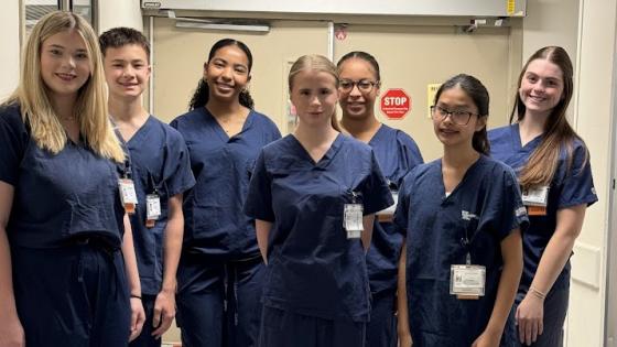 A group of high school students wearing their first pair of surgery scrubs, standing outside the surgery suite entrance during shadowing. 